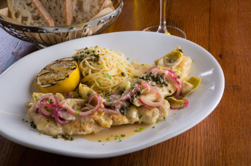 A plate of fish with lemon, capers, artichokes, and pasta, garnished with herbs, served with bread in the background.