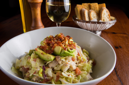 A bowl of salad topped with avocado and bacon, accompanied by a glass of white wine and a basket of bread.