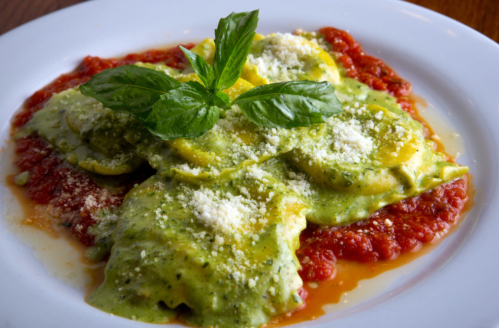 Plate of ravioli topped with green sauce, marinara, grated cheese, and fresh basil leaves.