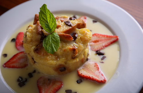 A dessert featuring bread pudding topped with mint, strawberries, and chocolate chips, served in a creamy sauce.