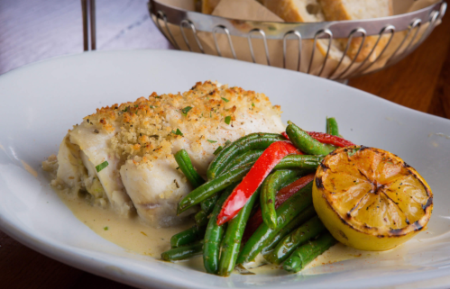 A plate of baked fish with a crispy topping, served with green beans, red peppers, and a grilled lemon half.