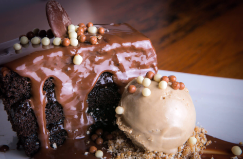 A slice of chocolate cake with dripping ganache, topped with colorful chocolate balls, alongside a scoop of caramel ice cream.