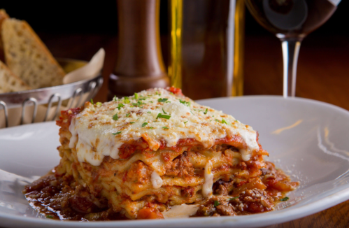A delicious serving of lasagna topped with cheese and herbs, accompanied by bread and a glass of red wine.