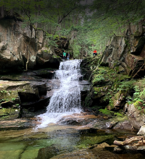 A serene waterfall cascades over rocks in a lush forest, with hikers visible on the rocky ledge above.