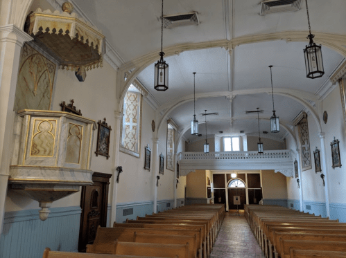 Interior of a church featuring wooden pews, decorative walls, and hanging lanterns, with a balcony on the left.