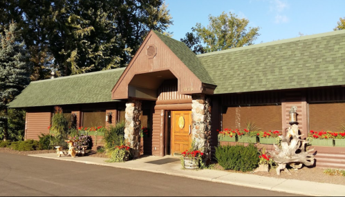 A rustic building with a green roof, stone pillars, and flower beds, set against a clear blue sky.