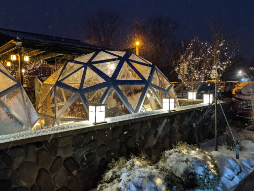 A cozy outdoor dining area with geodesic domes, illuminated by lanterns, surrounded by snow and festive lights at night.
