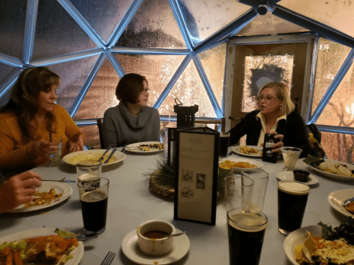 A group of women dining inside a geodesic dome, enjoying food and drinks with a foggy window in the background.