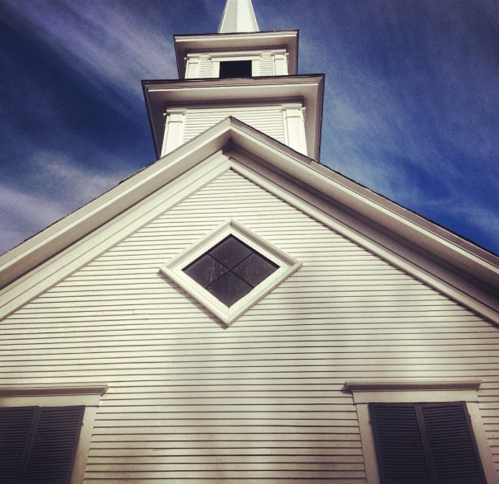A white church with a tall steeple and a diamond-shaped window against a blue sky.