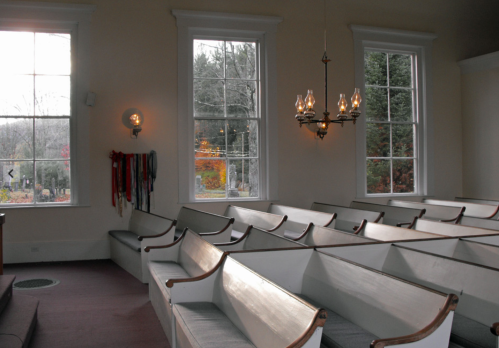 Interior of a church with wooden pews, large windows, and a chandelier, featuring a cozy, inviting atmosphere.