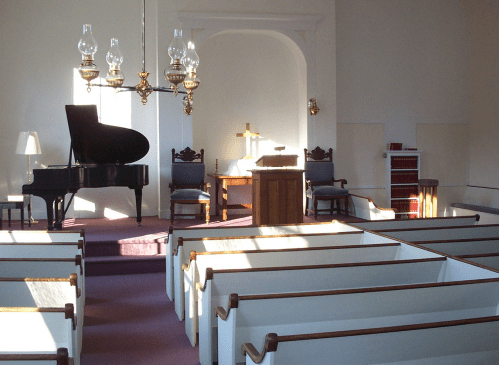 Interior of a church with a grand piano, pulpit, and pews, illuminated by natural light.