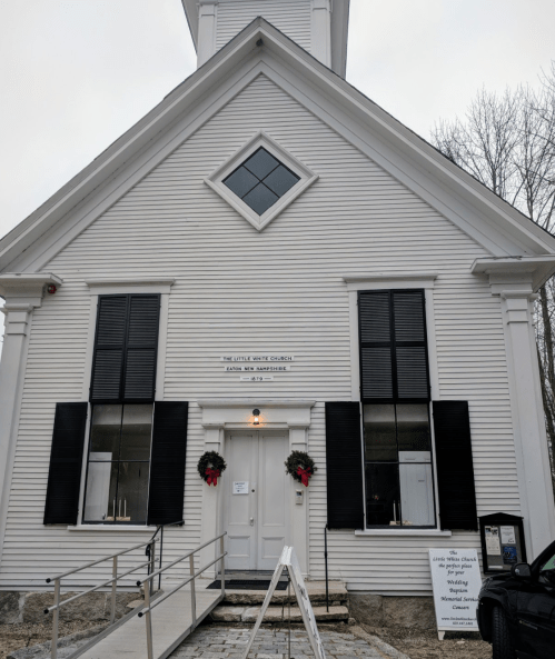 A small white church with black shutters, a steeple, and holiday wreaths on the doors. Signage is visible in front.