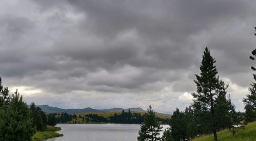 A serene lake surrounded by trees under a cloudy sky, with distant hills in the background.