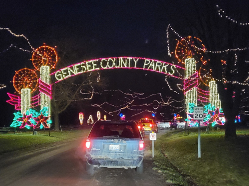 A car drives under a brightly lit archway reading "Genesee County Parks" adorned with festive decorations at night.