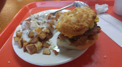 A plate with a biscuit sandwich filled with bacon and egg, alongside a serving of diced potatoes.
