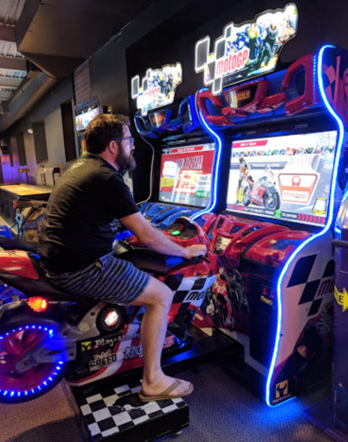 A person playing a motorcycle arcade game, sitting on a bike-shaped console with colorful lights in a gaming venue.