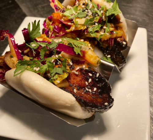 Two bao buns filled with grilled meat, colorful slaw, and garnished with herbs and sesame seeds on a white plate.