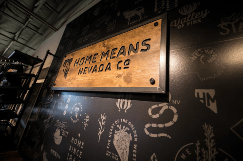 Wooden sign reading "Home Means Nevada Co." on a textured wall with various Nevada-themed graphics.