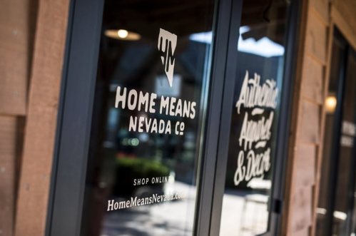Storefront window featuring "Home Means Nevada Co." logo and website, with a rustic wooden background.