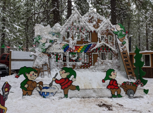 Colorful elf decorations around a snowy gingerbread house in a forest setting, with trees in the background.