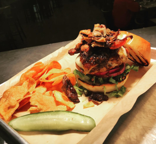 A gourmet burger topped with crispy onions, served with potato chips and a pickle on a tray.