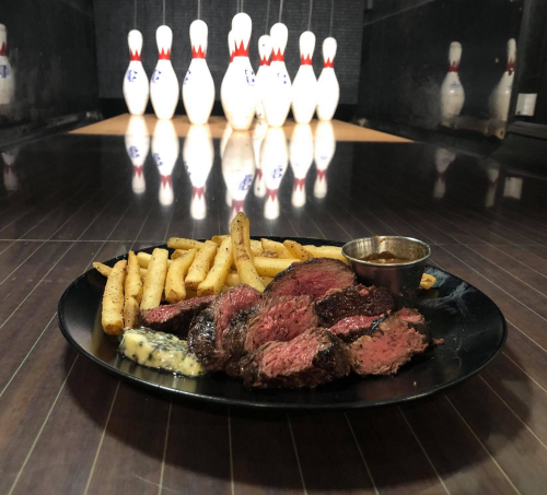 A plate of sliced steak with fries and dipping sauce, set against a bowling alley backdrop with pins in view.