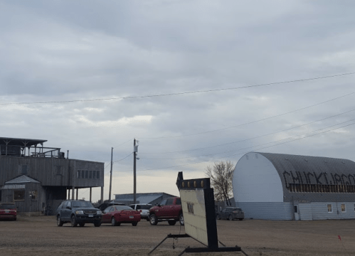 A rural scene featuring a wooden building, a large barn, and parked cars under a cloudy sky.