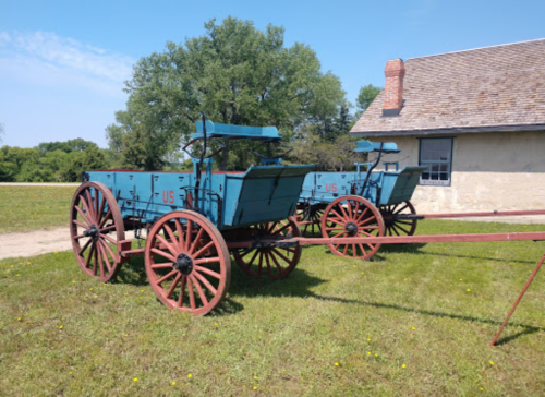 Two vintage blue wagons with red wheels parked on green grass near a building under a clear blue sky.