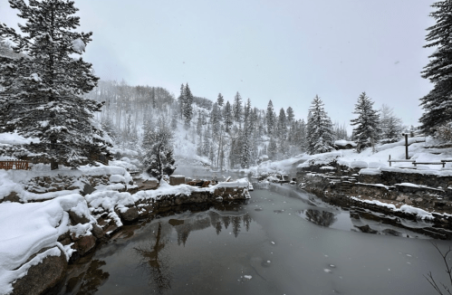 A serene winter landscape featuring a snowy riverbank surrounded by tall pine trees and mist rising from the water.