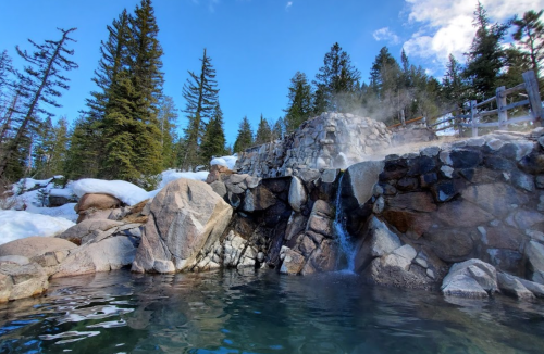 A natural hot spring surrounded by snow-covered rocks and evergreen trees under a clear blue sky.