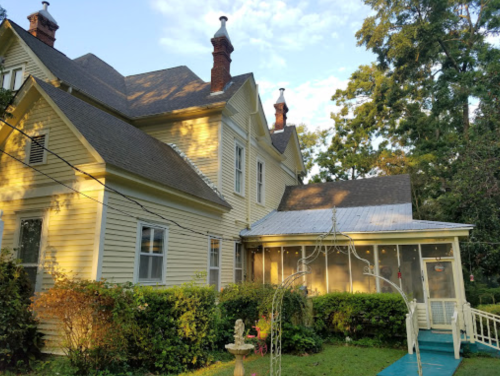 A large yellow Victorian-style house with multiple chimneys and a green lawn, surrounded by trees and shrubs.