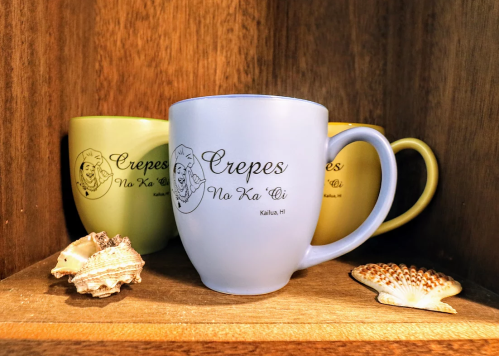 Three mugs on a wooden shelf, featuring the text "Crepes No Ka 'Oi" with seashells in front.