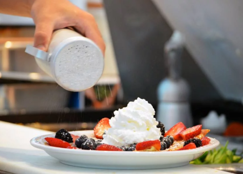 A hand sprinkles sugar over a dessert topped with whipped cream, strawberries, and blueberries on a plate.