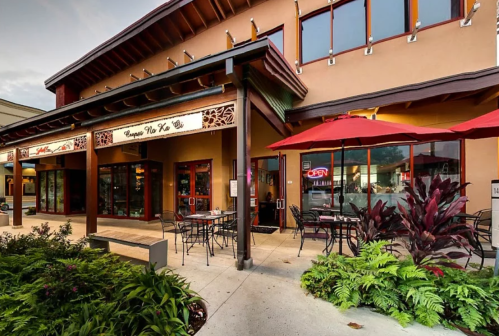 A restaurant exterior with outdoor seating, red umbrellas, and large windows, surrounded by greenery.