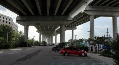 View of a parking lot under a highway overpass, with cars parked and greenery nearby.