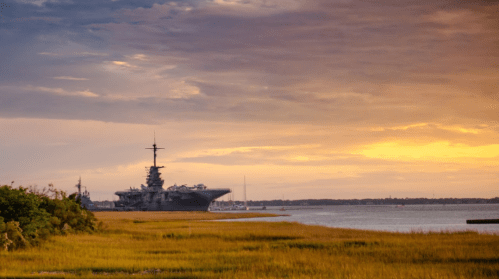 A naval ship docked near a grassy shore under a colorful sunset sky.