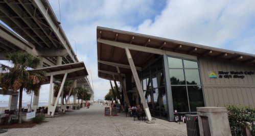 A modern café with large windows beside a walkway, palm trees, and a bridge overhead under a cloudy sky.