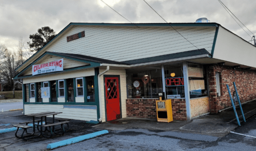 A small, brick restaurant with a red door and an "Open" sign, featuring large windows and outdoor seating.