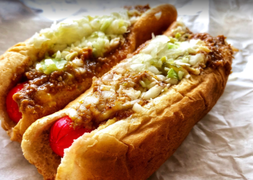 Two hot dogs in soft buns, topped with chili, chopped onions, and green onions, served on a paper background.