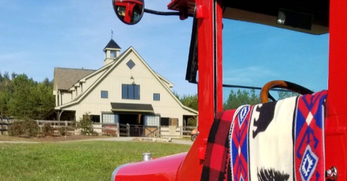 A red vintage truck with a blanket draped over the seat, set against a barn and green landscape.