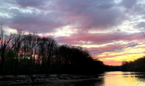 A serene river at sunset, with vibrant clouds and silhouetted trees along the banks.