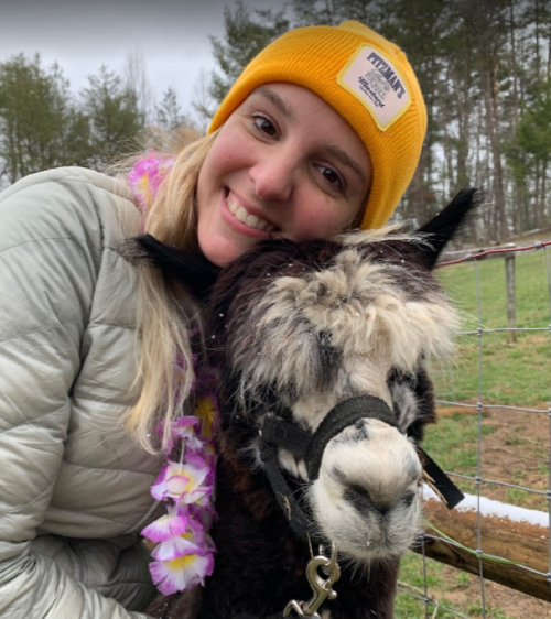 A smiling person in a yellow beanie hugs a fluffy alpaca, surrounded by greenery and a fence.