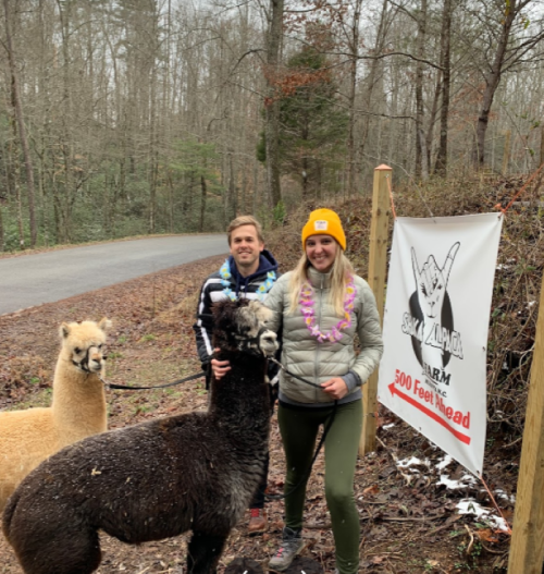 A man and woman stand with two alpacas near a sign for a farm, surrounded by trees on a cloudy day.