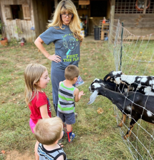 A woman and three children interact with goats at a farm, surrounded by greenery and a rustic barn in the background.