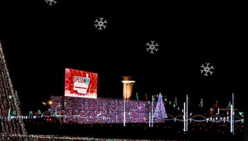 A festive display of colorful holiday lights with snowflakes in the night sky and a large sign in the background.