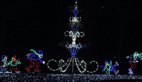 Colorful holiday lights depict children skating around a large, illuminated fountain in a dark setting.
