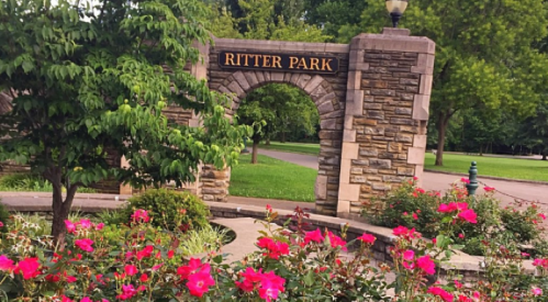 Stone archway with "Ritter Park" sign, surrounded by vibrant pink flowers and lush greenery.