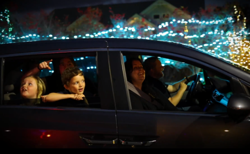 A family in a car enjoying a festive light display, with children smiling and pointing outside.