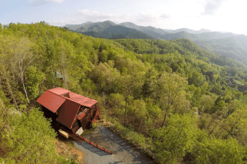 Aerial view of a rustic cabin surrounded by lush green trees and mountains in the background under a clear sky.