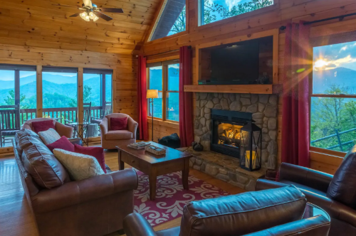 Cozy cabin living room with a stone fireplace, large windows, and mountain views, featuring comfortable seating and warm decor.